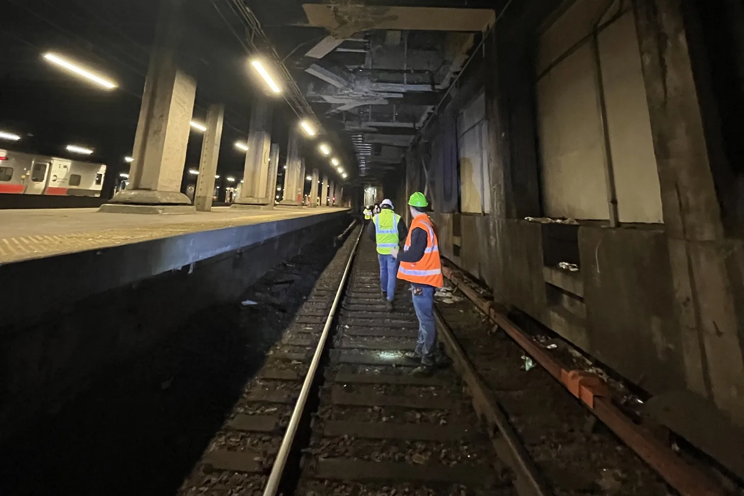 Two workers in safety vests and hard hats walking a track in an underground rail yard beside a commuter train, seen from behind